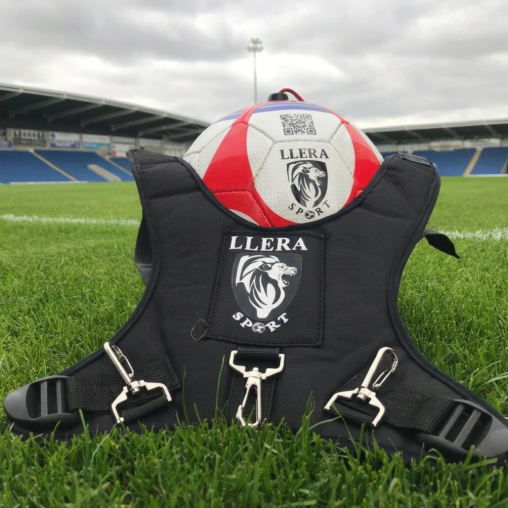 A black Llera Sport training harness laid on green grass at a soccer stadium, with a red, white, and blue soccer ball resting on top. The harness features silver metal clips and a black Llera Sport logo patch.